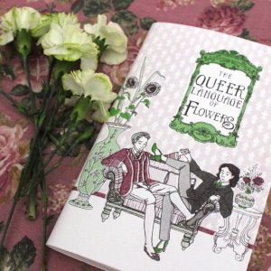 Image of a copy of The Queer Language of Flowers next to some green carnations. Two light-skinned people are sitting on a divan. One has short hair and is wearing a burgundy waistcoat, jacket, and breeches. The other has shoulder-length dark wavy hair and is wearing a green tie, a jacket and trousers, and green heels. There are sunflowers in a vase and roses and lavender in another.